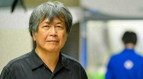 Portrait of Hsueh-Chia Chang wearing a dark button-up shirt, standing in a lab setting. A person in a blue lab coat is blurred in the background.