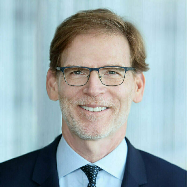 Smiling man with brown hair, goatee, and dark glasses wears a navy suit, light blue shirt, and patterned tie.